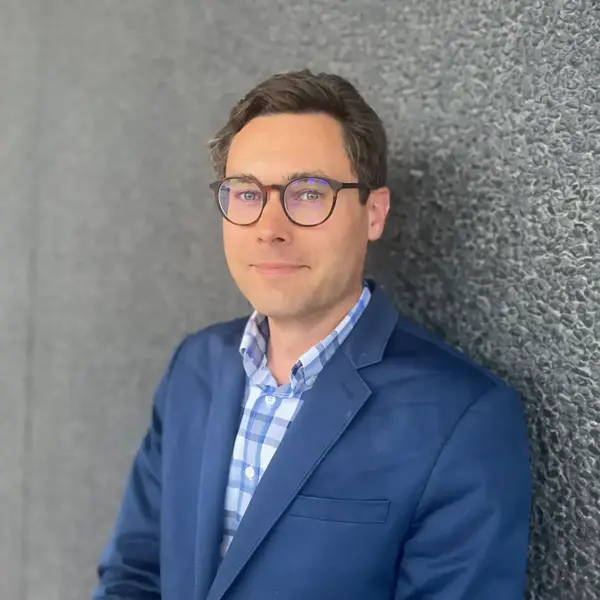 Gregory Laski, a man in a blue sports jacket and checkered shirt, wearing glasses, stands against a wall